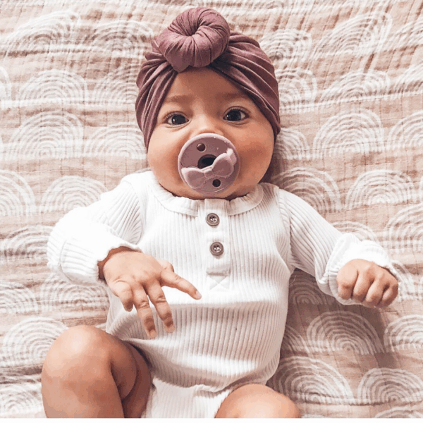 itzy ritzy sweetie soother™ pacifier sets (2 pack): agave + succulent cables Baby with pacifier and turban, wearing white outfit.