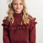 Girl in red sweater with ruffles smiling indoors.