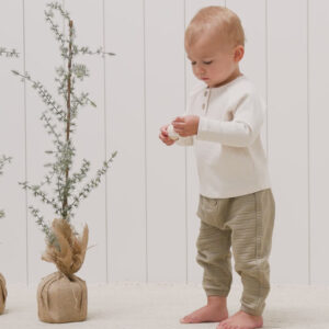 Toddler in beige outfit near small potted trees.
