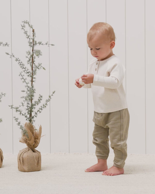 Toddler in beige outfit near small potted trees.