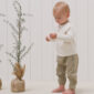 Toddler in beige outfit near small potted trees.
