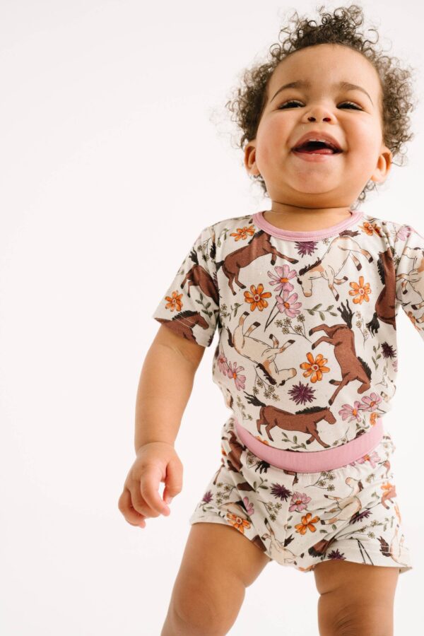 little one shop prairie ponies bamboo shorts set Smiling baby in floral horse-patterned outfit.