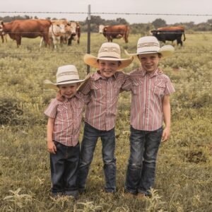Three kids in cowboy hats standing together near cows.