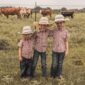 Three kids in cowboy hats standing together near cows.