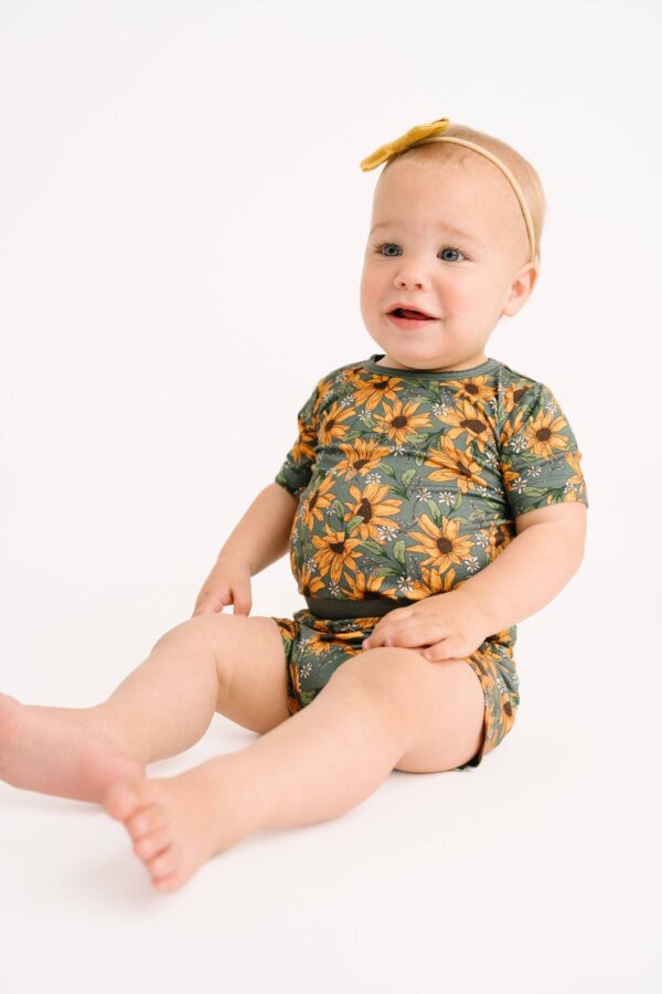 little one shop sunflower meadow bamboo shorts set Baby in floral romper with headband, sitting.