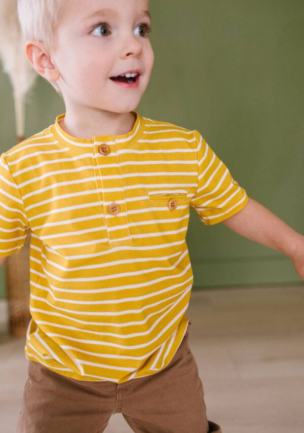 ollie jay alex tee in mustard stripe Smiling child in yellow striped shirt indoors.