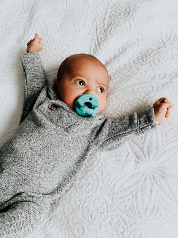 itzy ritzy sweetie soother™ pacifier sets (2 pack): agave + succulent cables Baby with pacifier lying on patterned blanket.