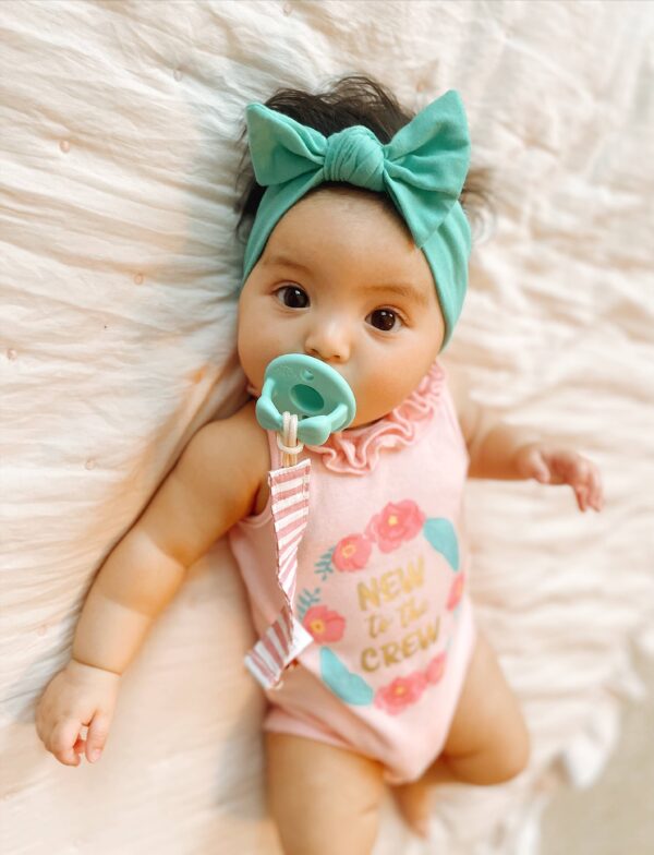 itzy ritzy sweetie soother™ pacifier sets (2 pack): aquamarine + peach bows Baby with pacifier and headband on blanket.