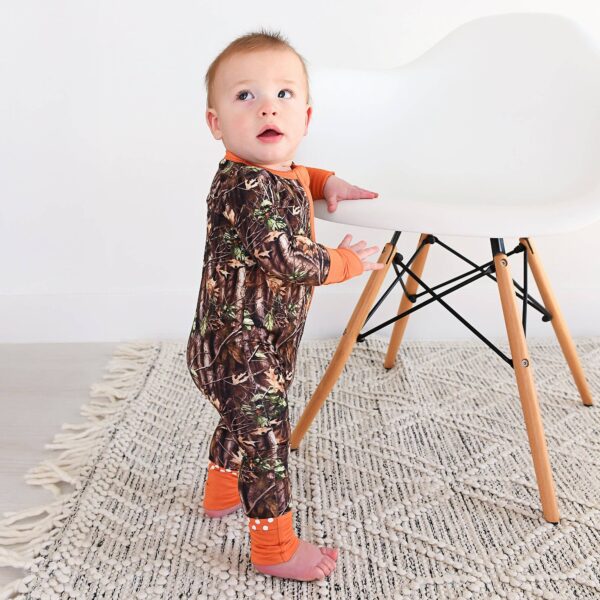 gigi and max hunter camo zip sleepies little bamboo baby outfit Baby in camouflage onesie stands by chair.