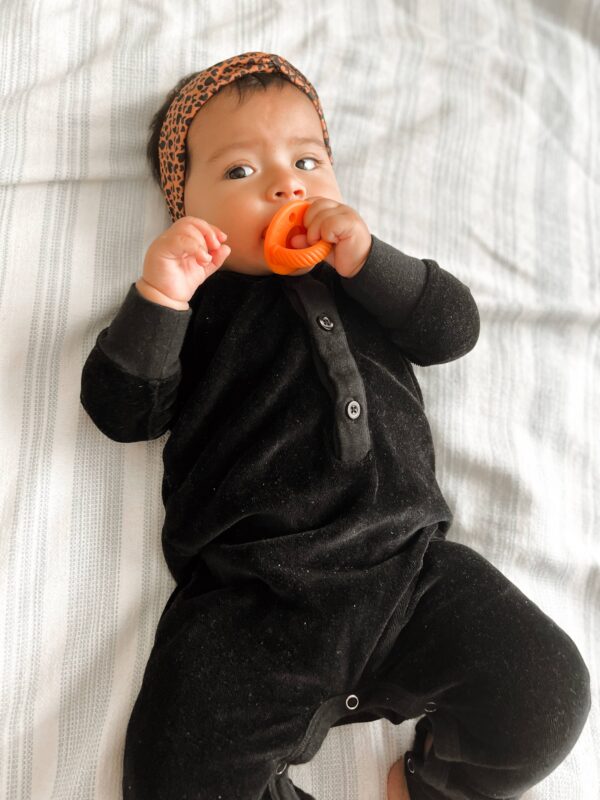 itzy ritzy sweetie soother™ pacifier sets (2 pack): aquamarine + peach bows Baby chewing toy on bed in black outfit.
