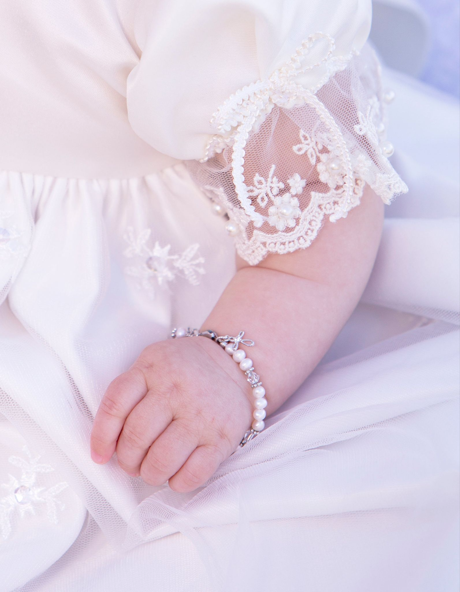 Baby hand wearing pearl bracelet, lace sleeve visible.